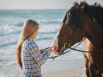 Young woman touching horse while standing against sea at beach