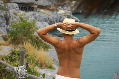 Rear view of shirtless young man with hands behind head standing by lake