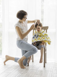 Mother cuts her son's hair by herself. little boy sits and holds pair of scissors. 