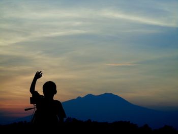 Silhouette boy with arm raised against sky during sunset