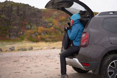 Side view of carefree female traveler in outerwear sitting in car trunk and enjoying hot drink from thermos cup while looking away