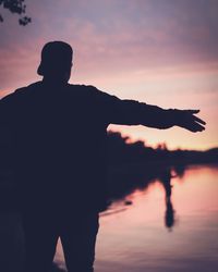 Silhouette man with arms outstretched standing at lakeshore against sky