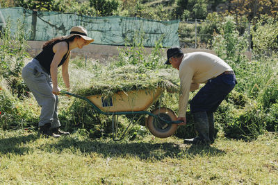 Mature farm worker helping colleague in lifting wheelbarrow at field