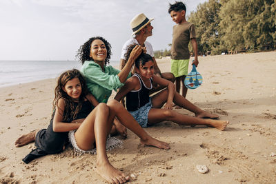 Portrait of smiling woman tying daughter's hair while sitting with family on sand at beach