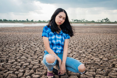 Full length of beautiful young woman crouching on crack land against sky