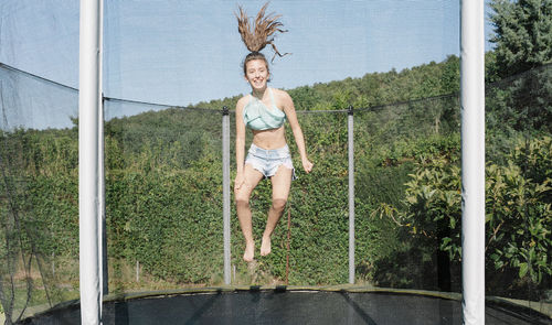 Horizontal photo of a smiling young brunette teenager jumping on a trampoline with net around on the green yard outdoors. the girl wears short jeans and a top and looks fun, active