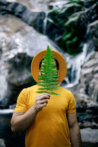 Peaceful male traveler in yellow clothes standing with green fern leaf on face and enjoying nature on background of waterfall in forest