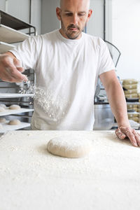 Male worker sprinkling white flour on unbaked dough ball on table while working in light professional bakery