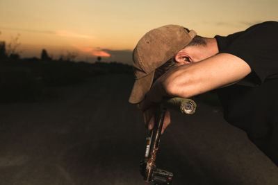 Side view of young man with bicycle sitting on road against sky during sunset