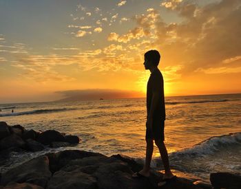 Silhouette teenage boy standing on rock at beach against sky during sunset