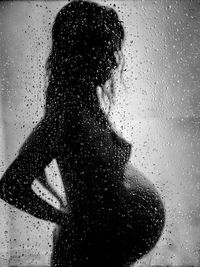 Silhouette pregnant woman standing in bathroom seen through wet glass