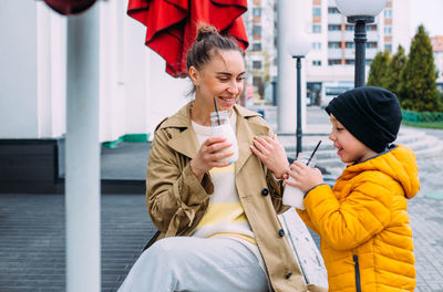 Young mom and son have fun and drink milkshake outdoors.