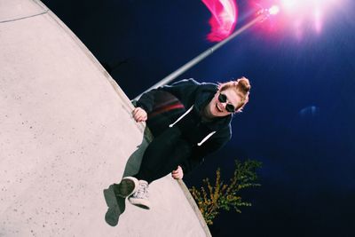 Tilt image of happy woman in sunglasses sitting on balcony wall at night