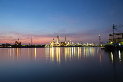 Illuminated factory by river against sky at dusk