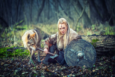 Portrait of young woman holding axe while sitting with wolf in forest