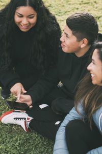 Three people of hispanic-latino ethnicity sitting on the green grass of a park, with smart device