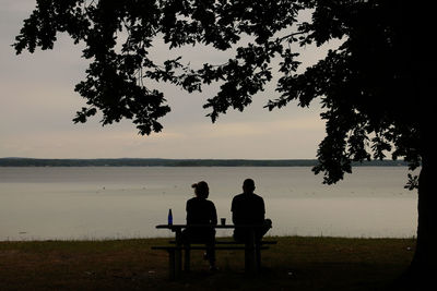 Silhouette men sitting on shore by sea against sky during sunset