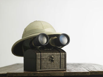 Close-up of binoculars on box with hat on table against white background