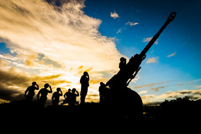 Silhouette soldiers saluting by cannon against sky during sunset