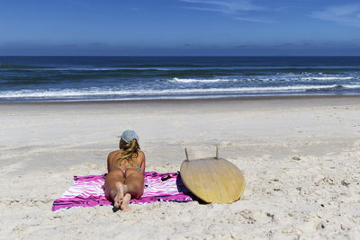 Rear view of woman in bikini lying by surfboard on blanket at beach against blue sky during sunny day