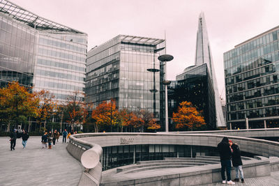 People walking by modern buildings in city against sky