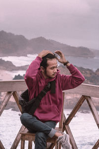 Young man sitting by sea against sky