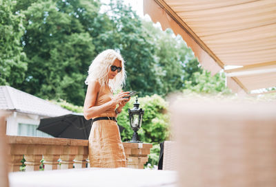 Adult elegant blonde woman in sunglasses and beige clothes using mobile phone in summer city cafe