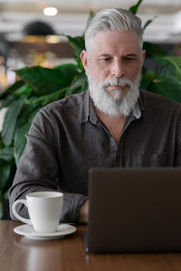 A grown man with gray hair and a beard works at his laptop in his office or airport waiting area