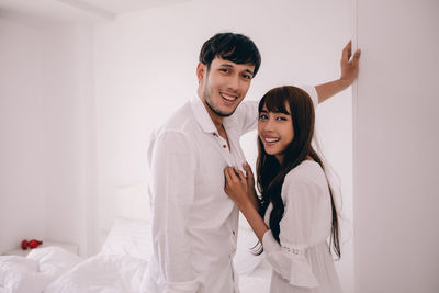 Portrait of smiling young couple standing in bedroom against wall at home