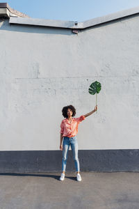 Smiling woman with hand raised holding monstera leaf in front of wall