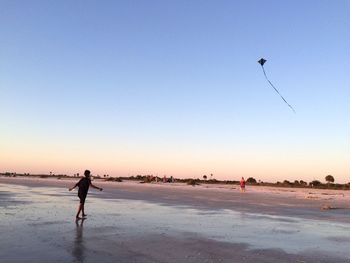Man walking at beach against kite flying in clear sky during sunset