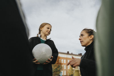 Female coach giving advice to elementary girl with sports ball against sky