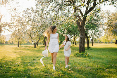 Mom and her daughter in white dresses run through a blooming apple orchard in spring on a sunny day 