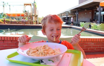 The blond boy closes his eyes and smiles. a five-year-old boy eats pasta and holds a fork and knife.