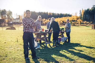 Portrait of mature female farmer standing arms crossed with friends selling food in background at field