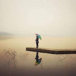 Side view of woman with umbrella standing by lake against sky