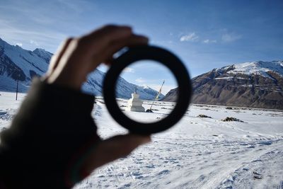 Cropped hand holding lens against built structure on snowcapped mountain