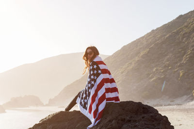 Portrait of woman wrapped in american flag sitting on rock at beach