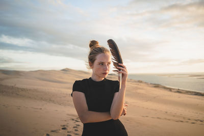 Serious female with black feather wearing dress standing on sandy dune washed by sea at sundown