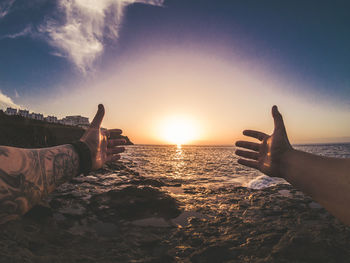 Cropped hands of man reaching sea against sky during sunset