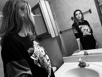 Black and white portrait of young woman standing in bathroom looking in a mirror.