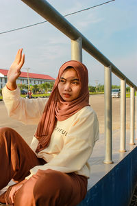 Portrait of young woman sitting on railing against sky