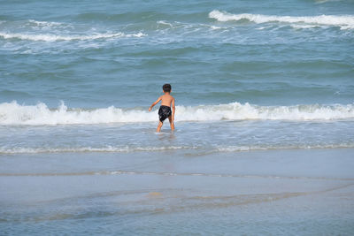 Rear view of shirtless boy standing by waves at beach