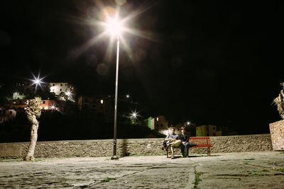 People on illuminated street light against sky at night