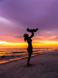 Young adult woman mother holding baby in air on beach with sunset background silhouette