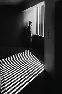 Side view of young man looking through window while standing in corridor