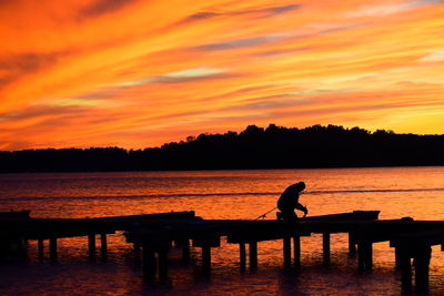 Silhouette man on pier by lake against orange sky