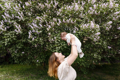Mother picking up baby girl by flowering plant at park