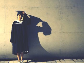 Student wearing graduation gown and mortarboard while standing against wall