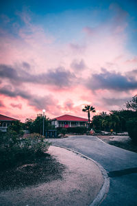 Road by buildings against sky during sunset